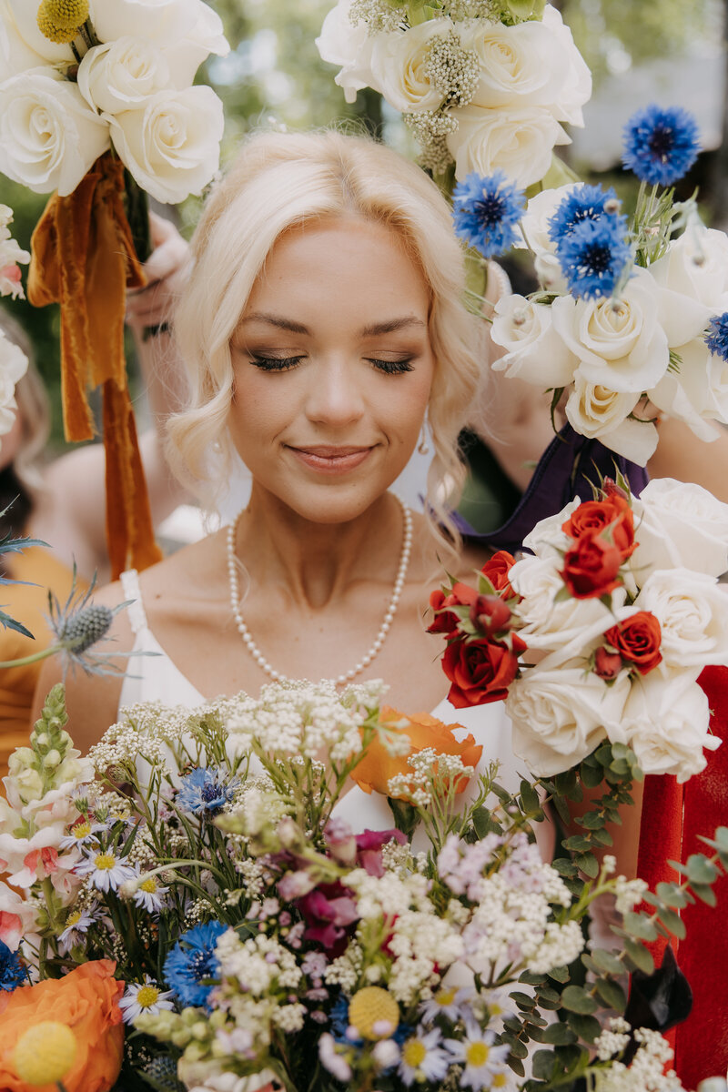 Bride holding a vibrant fall-colored bouquet, standing in the grass before a serene pond at sunset, bathed in golden light.