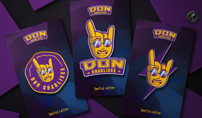 An array of enamel pins with three designs on the forefront: rock hand symbol with a smiling face wearing reflective sunglasses. The brand name is Don Charlieee.