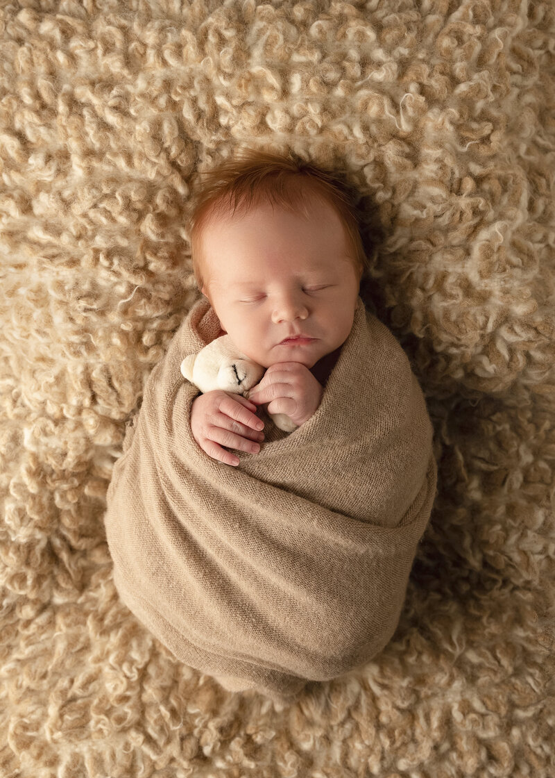 nPeaceful newborn baby boy’s first cuddles with teddy bear with spiky red hair, cozily swaddled on a tan blanket. Captured in Fort Mill SC by Insley Photography