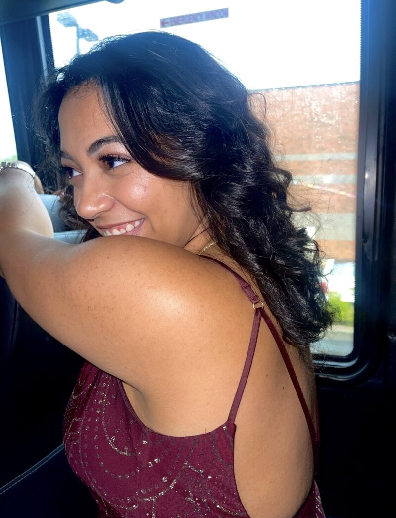 A woman in a maroon dress looking away while smilling and grabbing on to the seat in front of her.