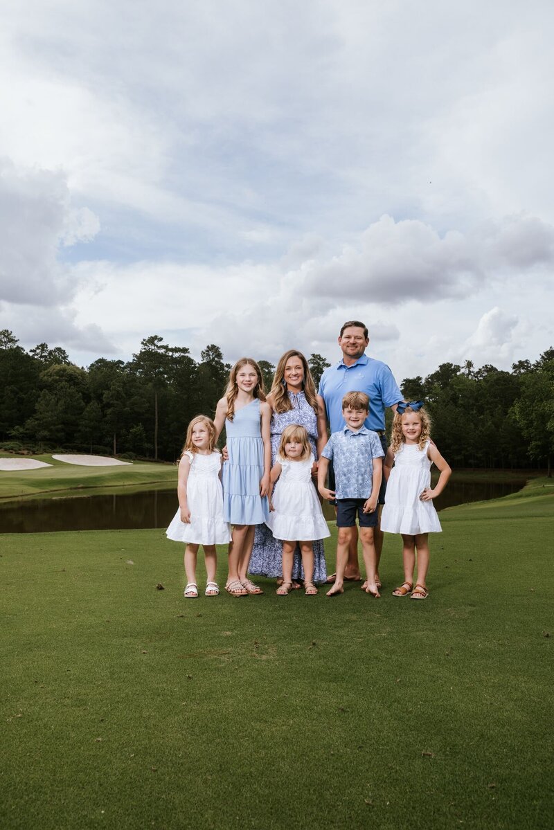 Large family with five children posed in coordinated outfits on a green golf course under cloudy skies in Tomball, Texas