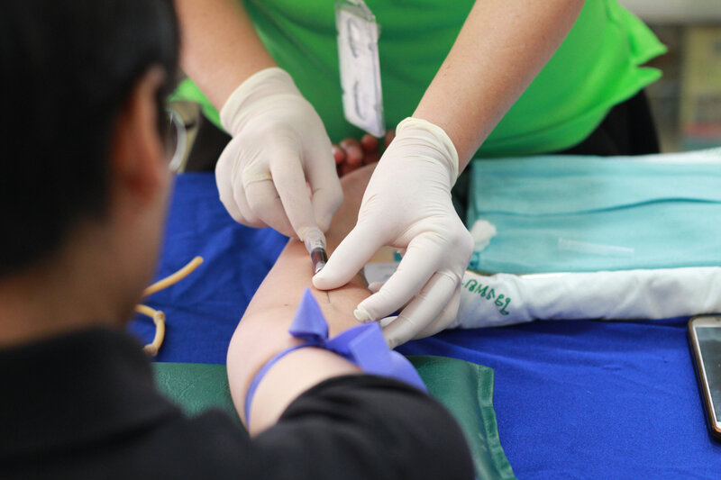 Healthcare worker wearing gloves drawing blood from a patient’s arm with a needle and tourniquet.