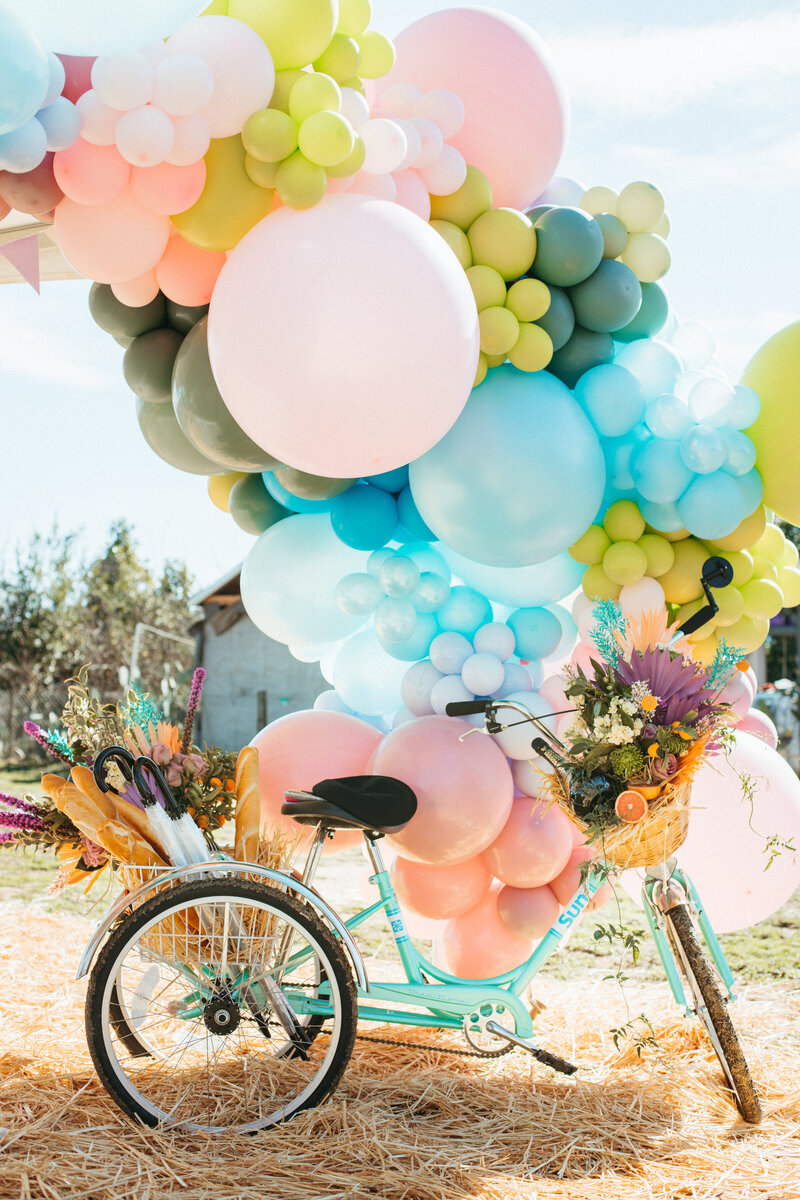 A whimsical scene featuring a mint-green tricycle decorated with vibrant spring flowers, baguettes, and umbrellas in its baskets. Above it floats a massive pastel balloon garland in shades of pink, yellow, green, and blue. Set outdoors with hay underfoot, this cheerful setup evokes a retro, carefree vibe perfect for a styled event or outdoor festival. Captured by Dani Lacey Photographs.