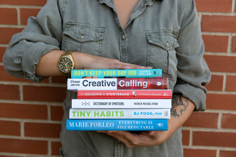 Calgary brand storytelling photoshoot of a person wearing an olive-green button-up shirt holding a neatly stacked pile of six books against a red brick wall. The book titles focus on creativity, personal growth, and business, including ‘Don’t Keep Your Day Job’ by Cathy Heller, ‘Creative Calling’ by Chase Jarvis, ‘Building a StoryBrand’ by Donald Miller, ‘Dictionary of Emotions’ by Patrick Michael Ryan, ‘Tiny Habits’ by BJ Fogg, and ‘Everything is Figureoutable’ by Marie Forleo. A gold watch and a tattoo on their forearm are visible.