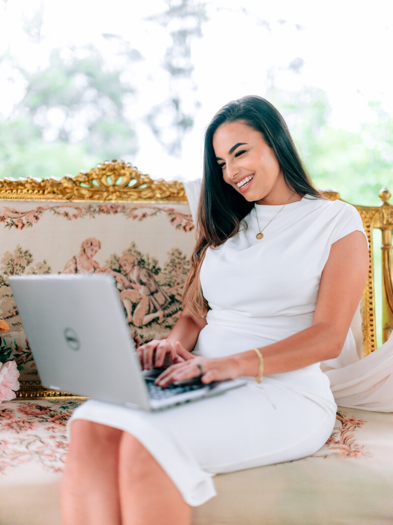 Event planner Linda Linares smiling while typing on a laptop on an ornate gold sofa.