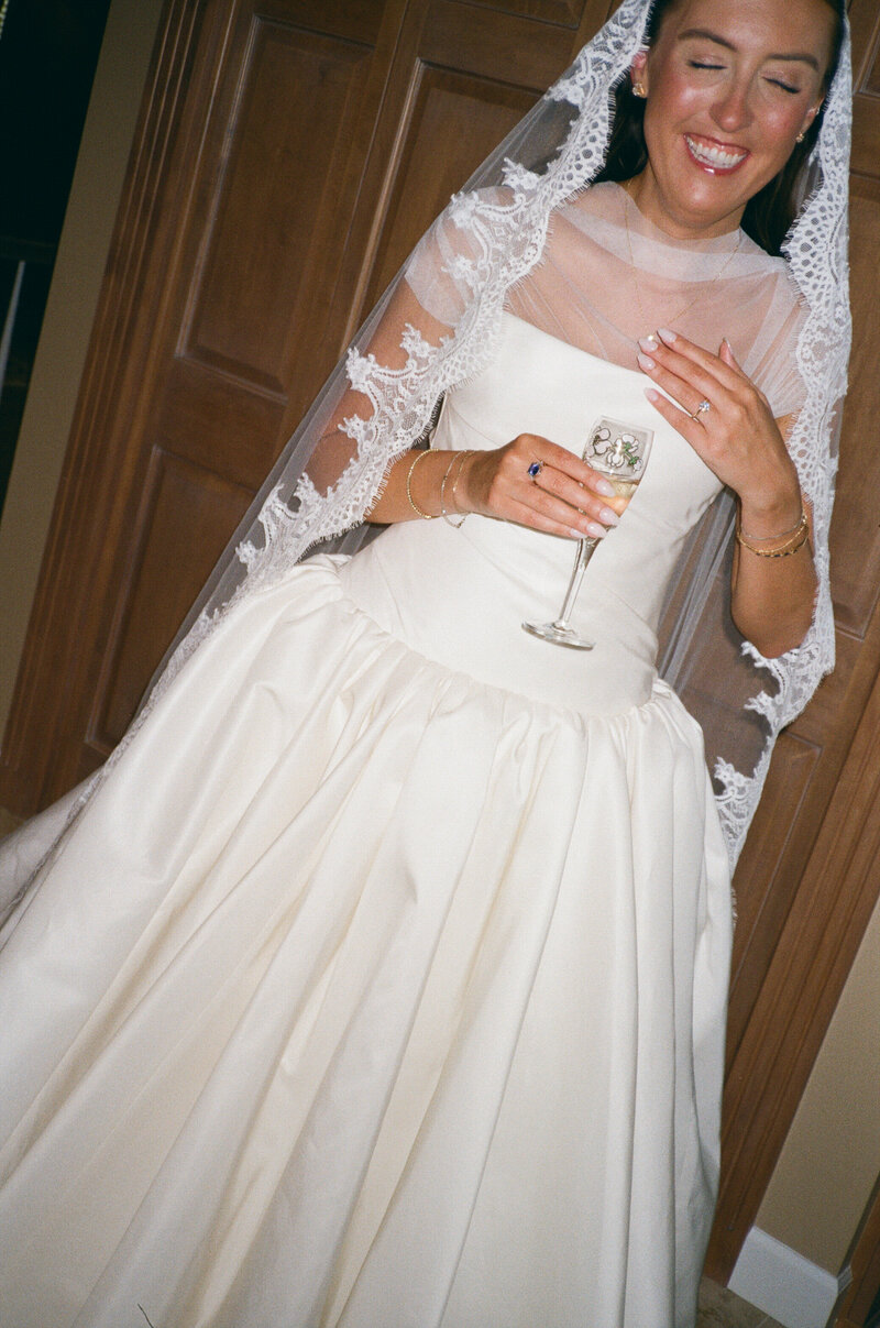 Bride in vintage-inspired satin wedding gown holding champagne glass and showing lace veil