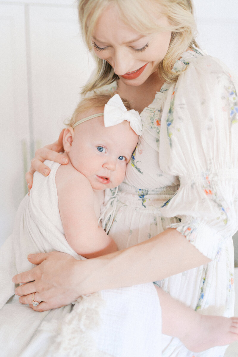close up of mom in a white floral dress sitting and holding her one year old daughter wrapped in a white towel with a white headband