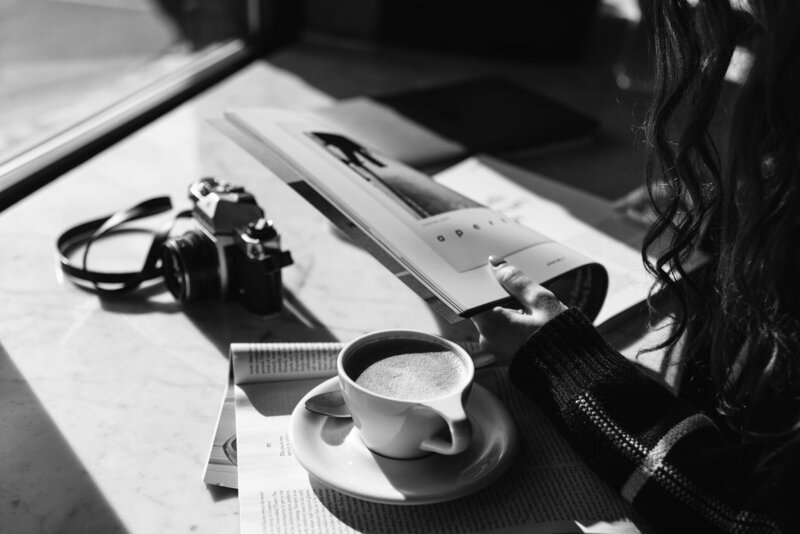 Black and white photo of woman reading a magazine at a cafe with a camera and latte next to her