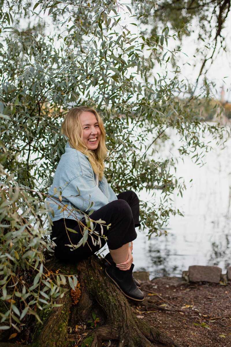 mielen paremminvoinnin ja kehomieliyhteyden valmentaja istuu rannalla puun alla ja hymyilee kameralle