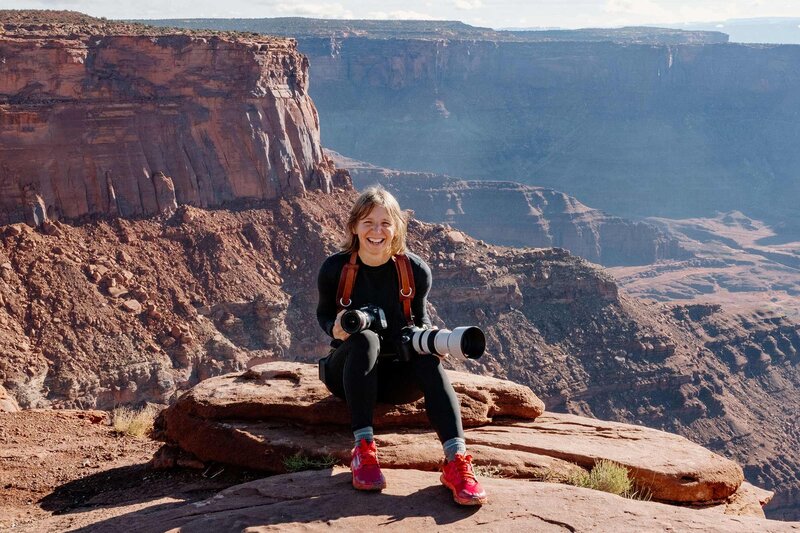 Aimee holds two cameras on top of a rock with mountains behind her.
