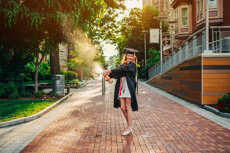 Female college graduate in a cap and gown spraying champagne into the air on the Temple University campus with the sun behind her