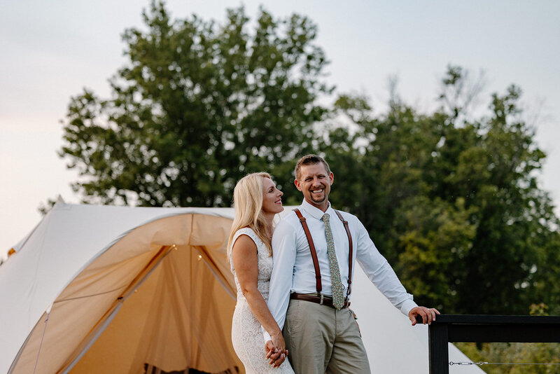 A bride looks at her husband smiling on their elopement day. They are holding hands and standing in front of a glamping tent
