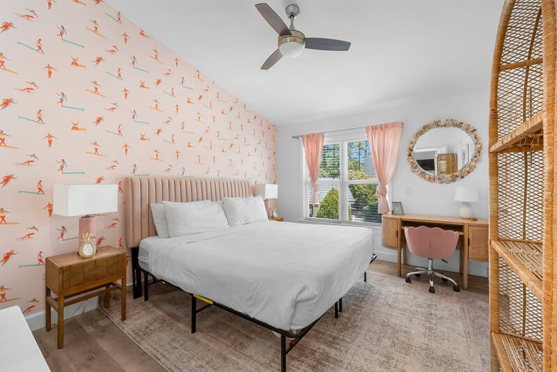 Primary bedroom with king-size bed, playful pink surfer wallpaper, blush curtains, and a coastal desk nook with seashell mirror.