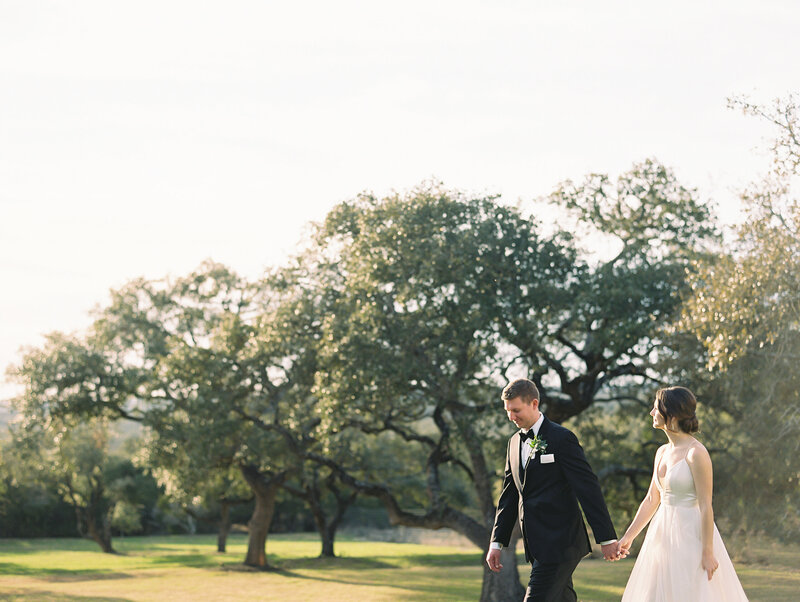 Bride and groom holding hands walking across the grass with trees in the background