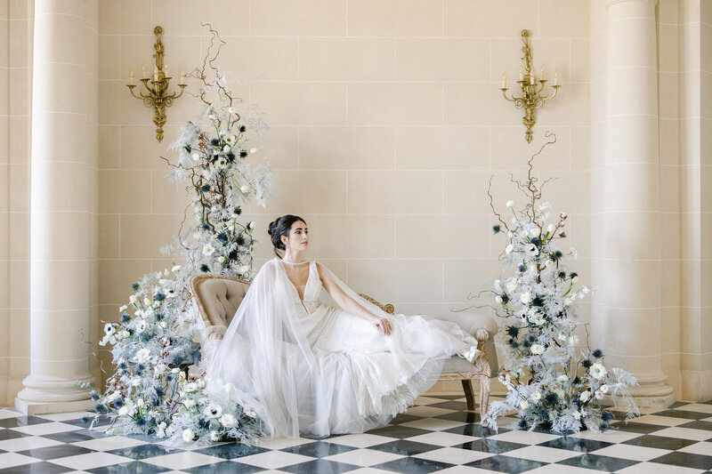 Mariée assise entre deux compositions florales dans le hall à colonnes du Château de Nainville, photographiée par Marion Pinel