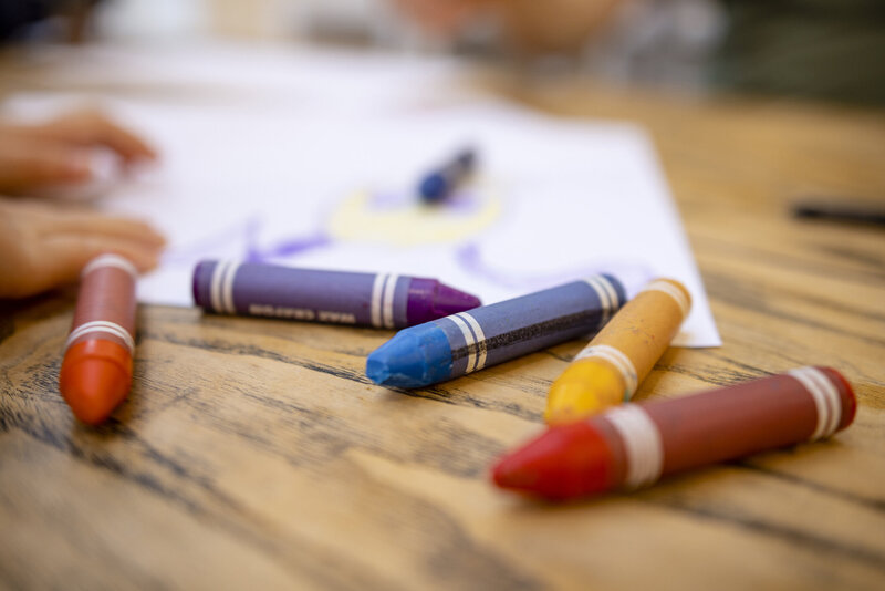 A close view of colorful crayons on a table with a child's drawing in the background