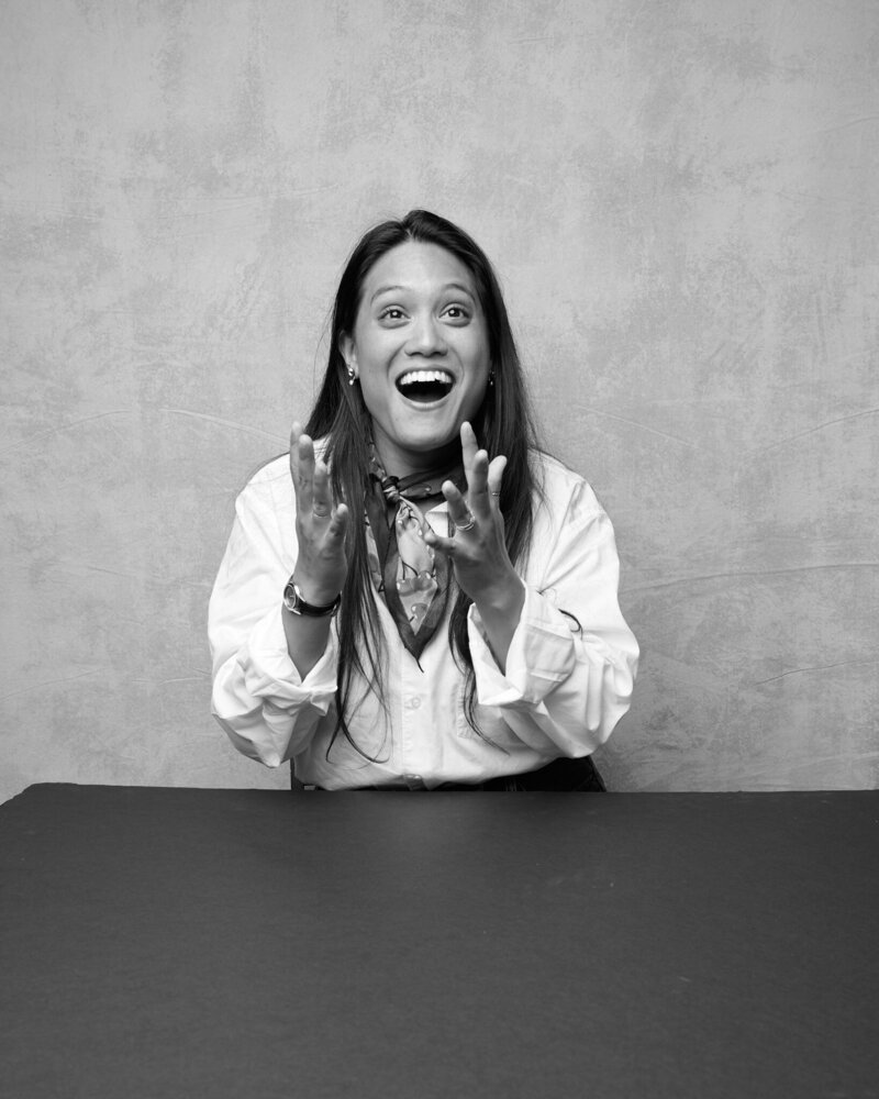 Black and white portrait of a woman sitting at a table, mid-gesture with wide eyes and an excited, joyful expression. She wears a white button-up shirt, a stylish scarf, and expressive jewelry, radiating energy and enthusiasm. A lively, unfiltered moment from 'THIS IS ME – A Photography Experience by Katie Campbell' in Atlanta, GA, celebrating personality and authentic self-expression.
