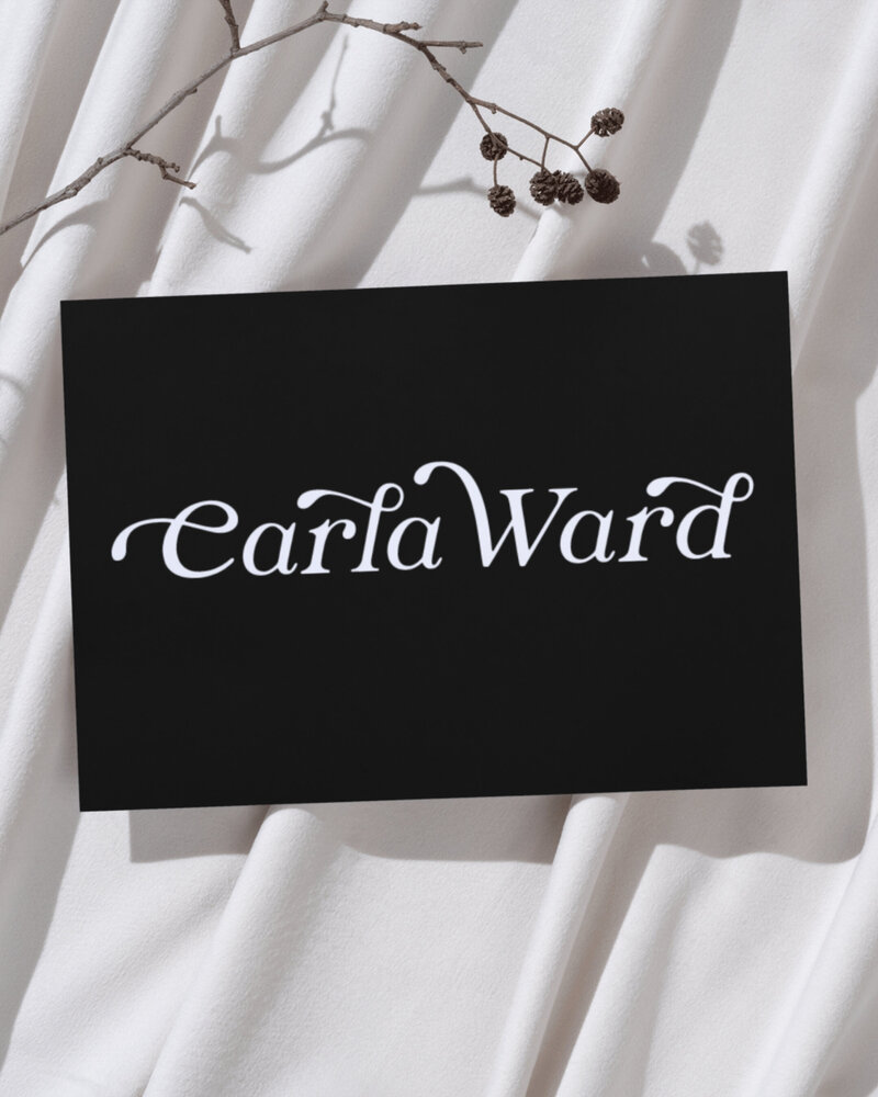 Elegant black card with the white Carla Ward logo displayed on cream fabric with natural light and a branch casting shadows.