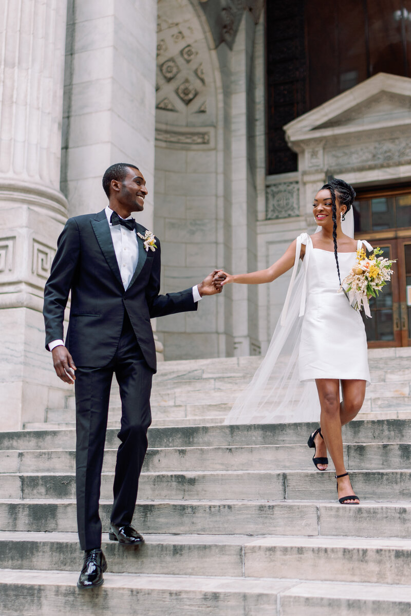 wedding couple ascends the stairs of the new york public library in alexandra grecco dress