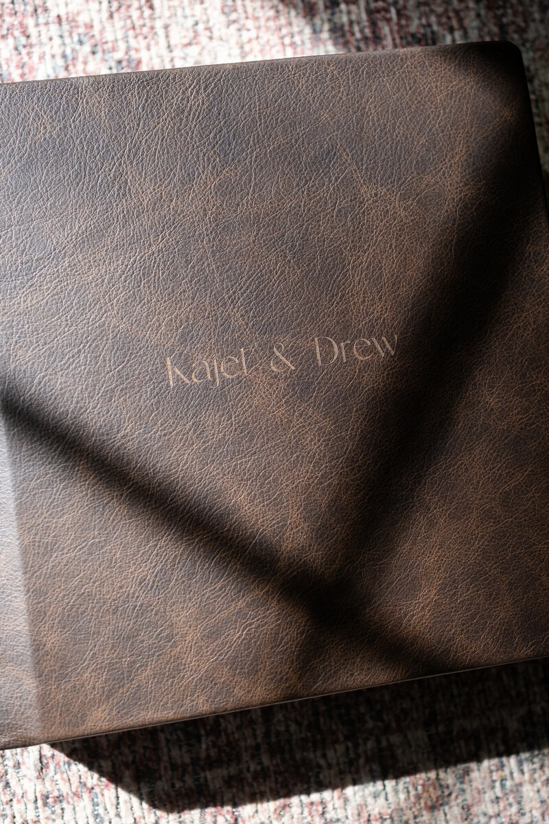 A luxurious, brown leather wedding album with elegant embossed names rests on a textured rug, illuminated by natural sunlight. This heirloom album, crafted to preserve a couple’s love story, showcases the timeless quality of Alabama wedding photography.
