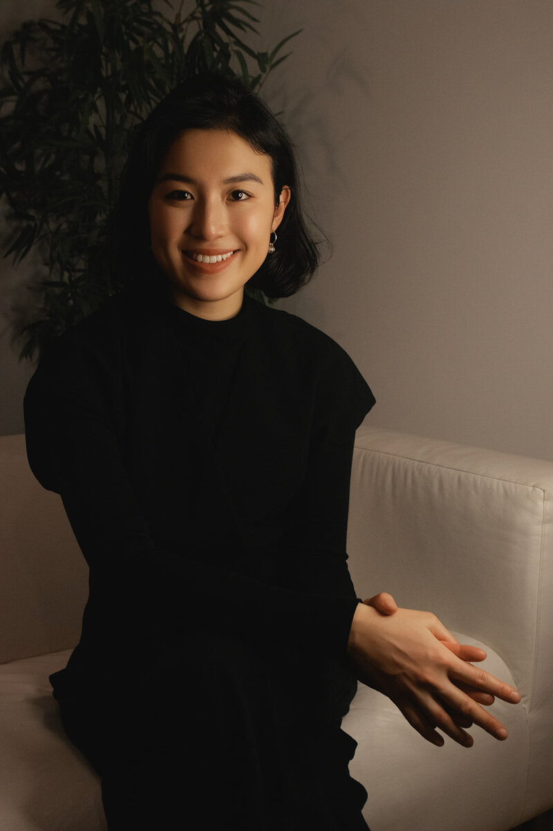 Woman with short dark hair weating a black long-sleeved shirt smiles at camera while sitting on an off-white couch with a tree behind her.