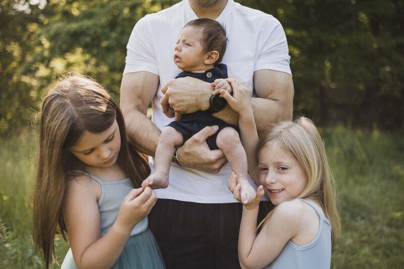 Dad holding baby while sisters touch baby's toes