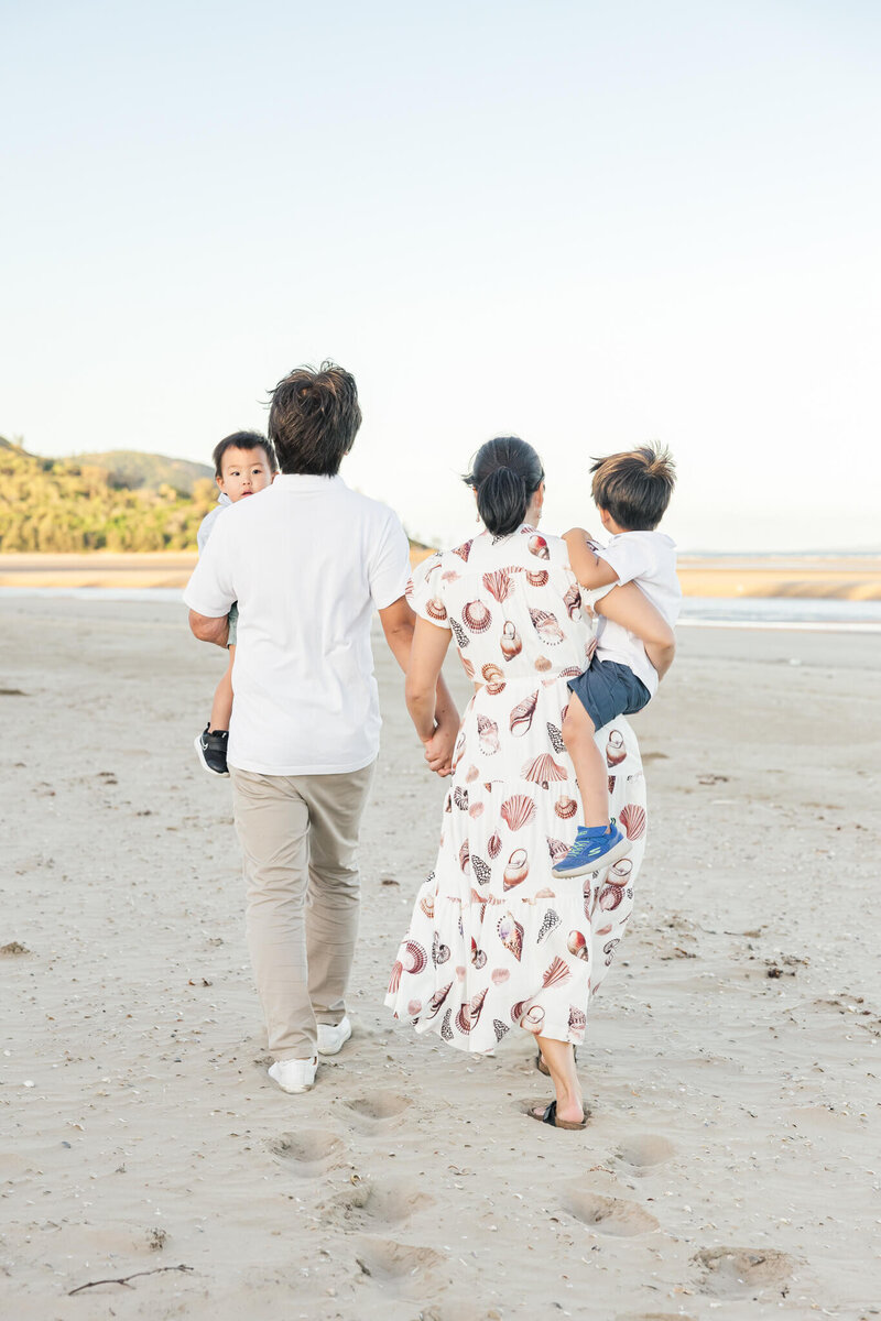 Stylish family walking along the beach, while Dad and Mum hold hands and hold their little boys in their arms.