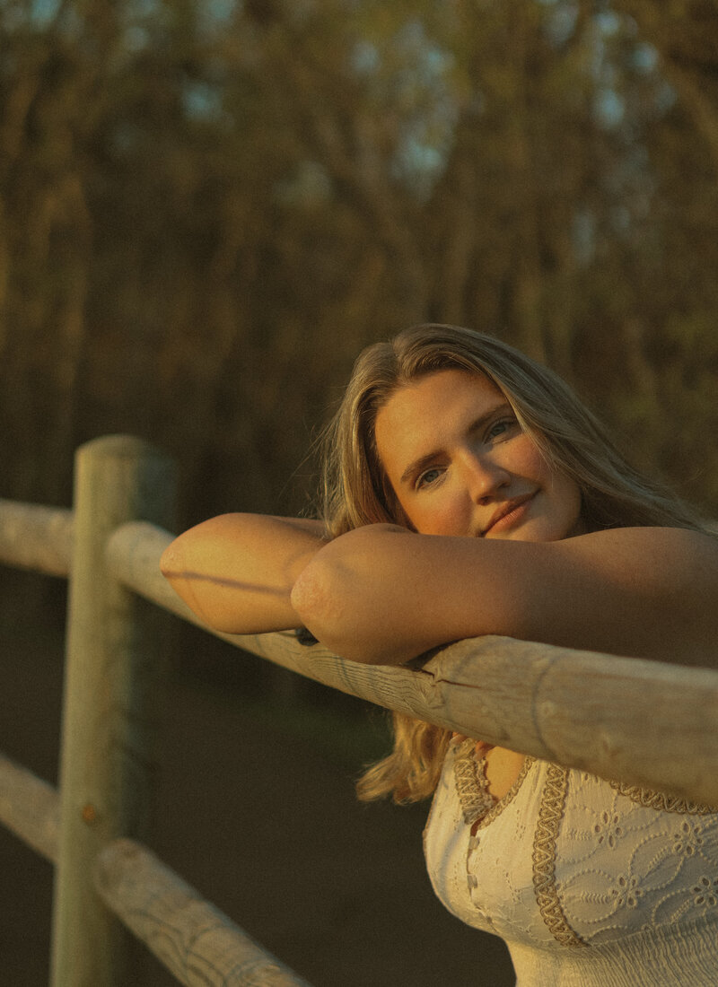 A portrait of a woman. her arms are crossed and resting on a wooden fence. Her head is resting on her arms and she has a soft smile while looking at the camera.