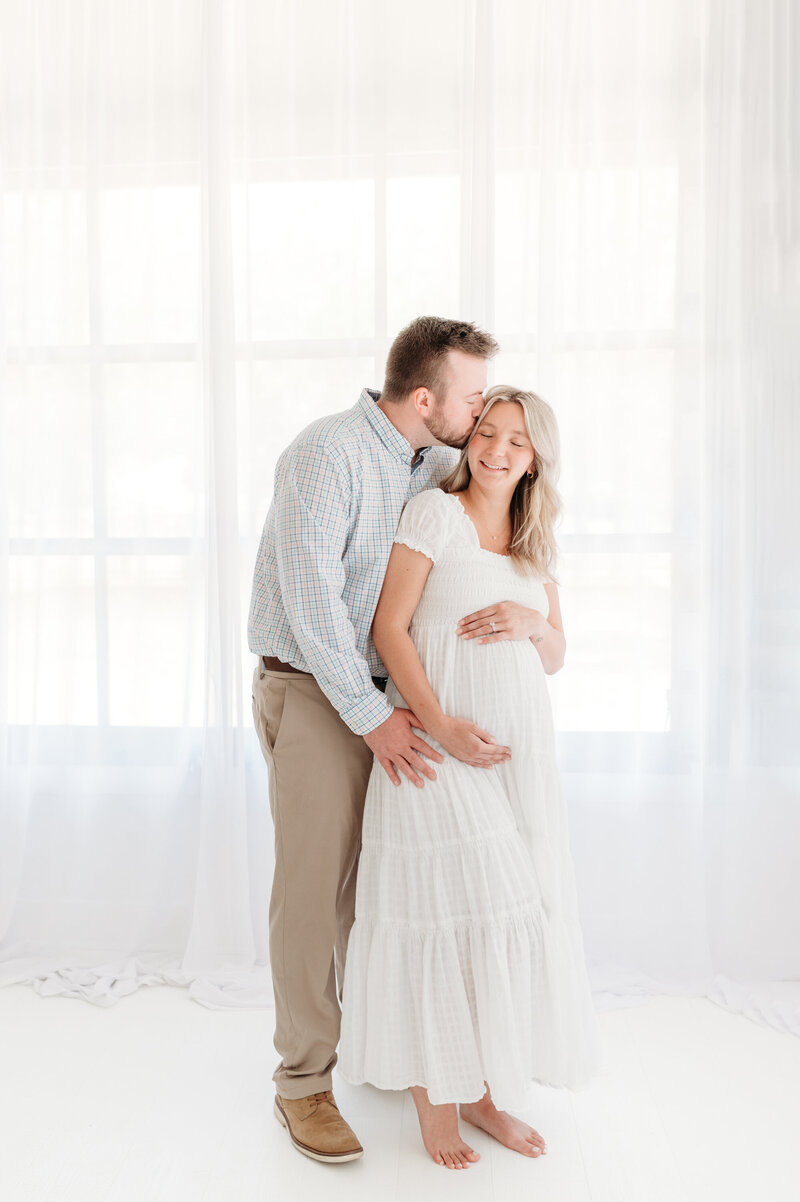 Dad is kissing and holding his arms around his pregnant wife, while she is smiling. Maternity photos taken in studio in Atlanta.