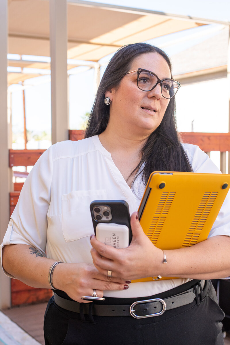 Professional woman holding a yellow laptop and phone while standing outdoors under a shaded patio, photographed by Vyrl Photo for Tucson brand photography.