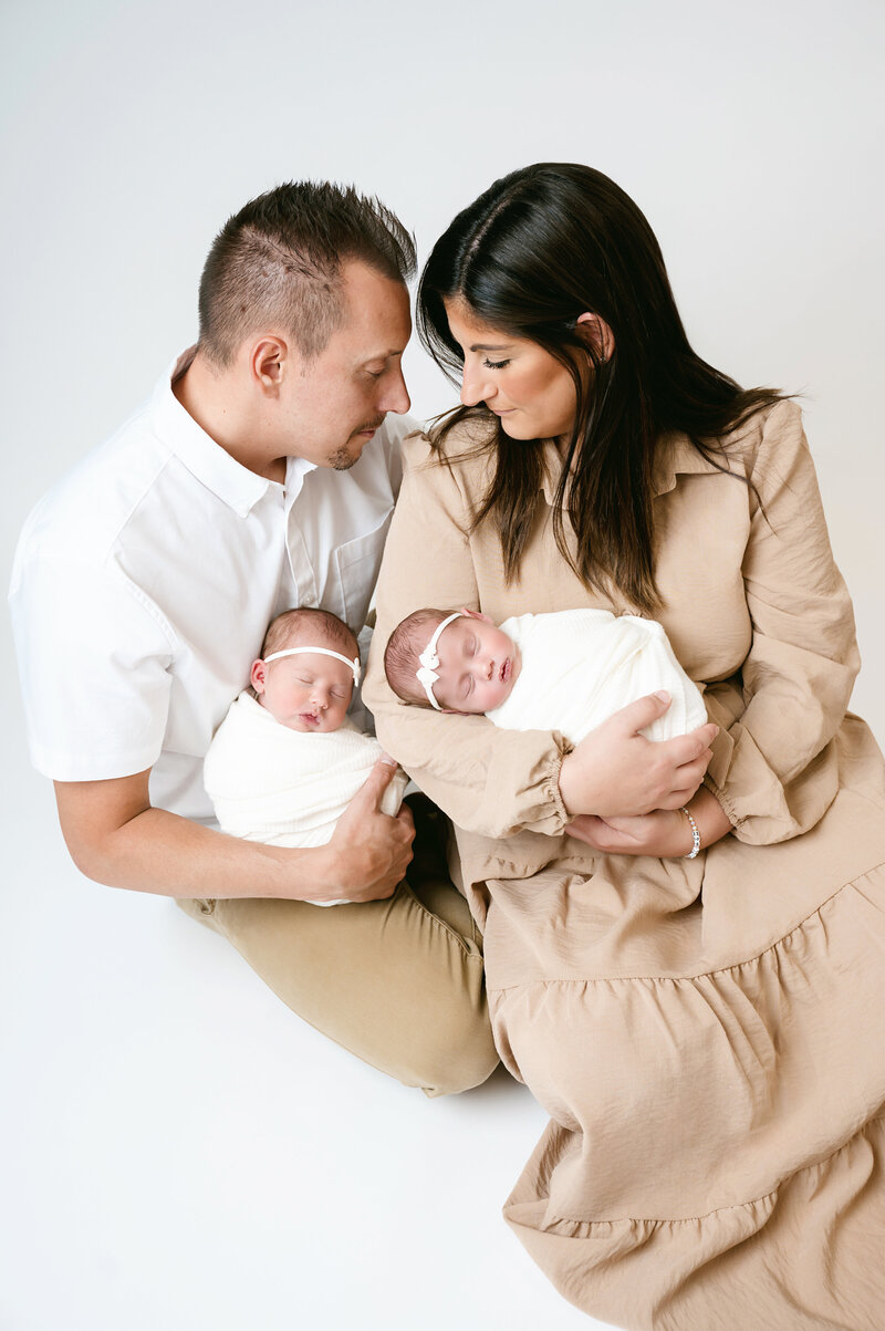 A beautiful family portrait with newborn twins, captured in a professional photography session in Pittsburgh. The loving parents hold their precious babies swaddled in soft white blankets, showcasing the joy and warmth of a growing family.