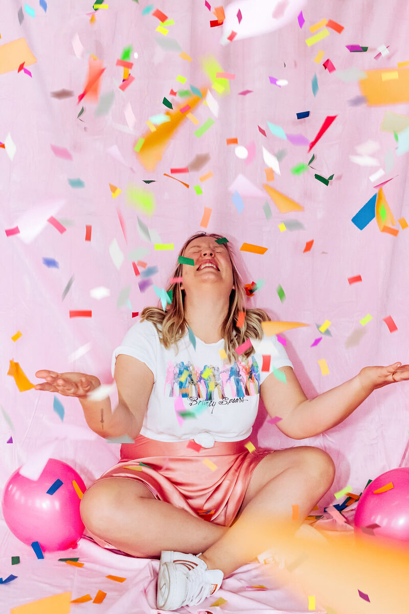 Jenn is laughing, sitting on the floor cross-legged as confetti falls down around her.