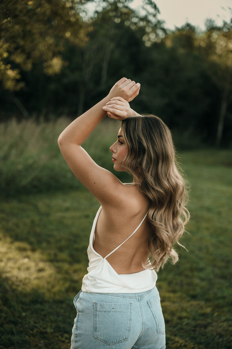 A woman in a white strappy top and blue jeans stands in a grassy field. She faces to the side, arms raised gracefully, exuding a serene and contemplative mood.