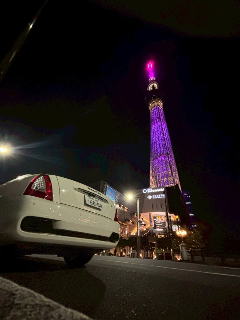 A white Maserati parked under the glowing Tokyo Skytree at night, illuminated in purple against the dark sky.