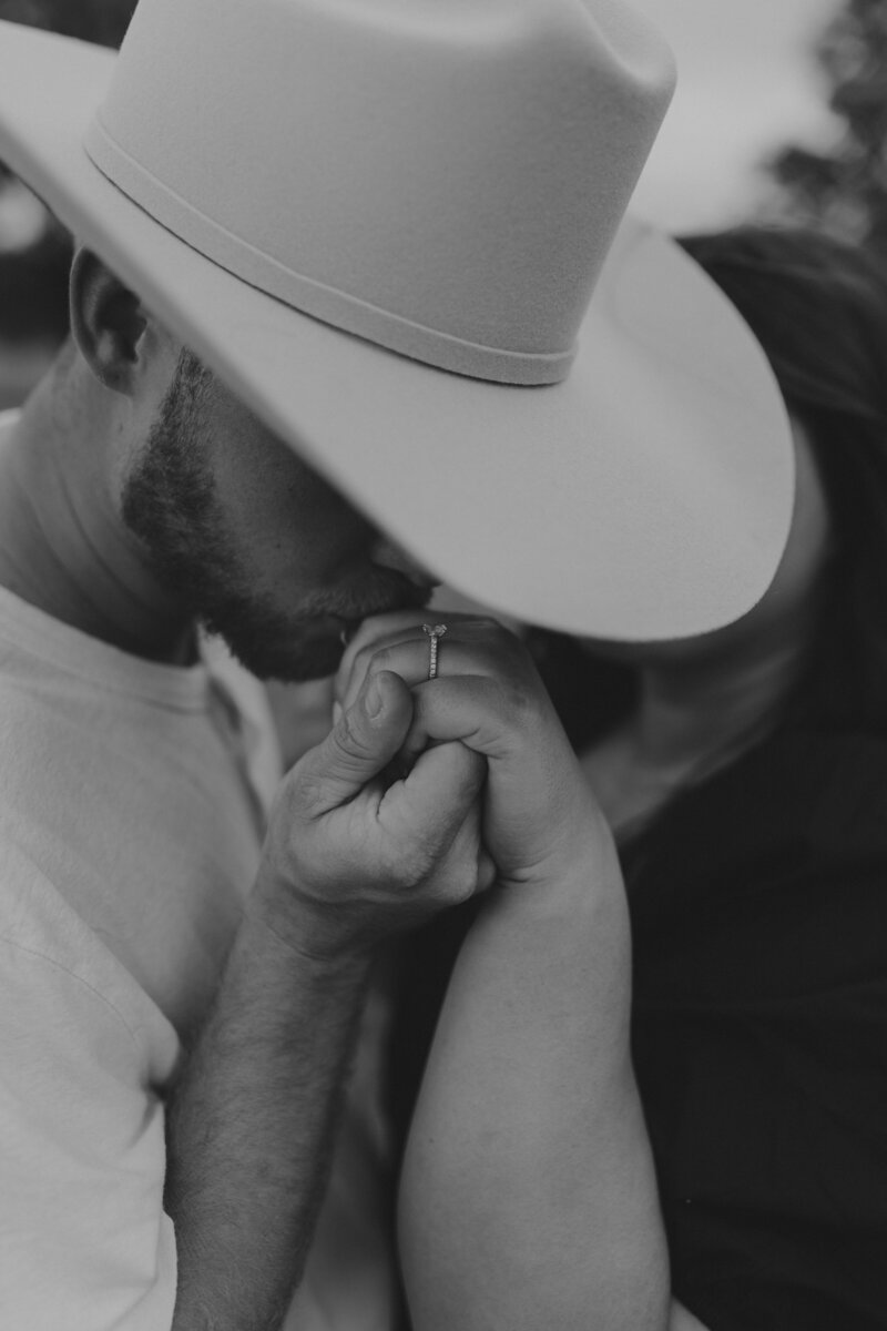 Engagement photo of a man wearing a cowboy hat kissing the love of his life hand and showing her engagement ring 