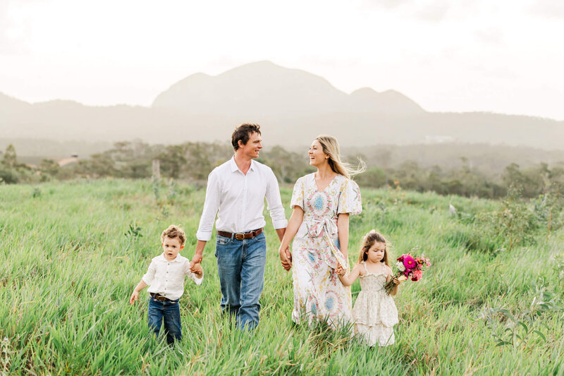 Stylishly dressed family of four walking through green grass with a mountain range as a backdrop. Dad and Mum holding hands of their son and daughter while they glance happily at each other.