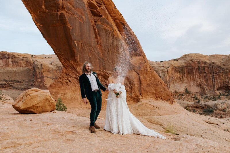 A bride and groom pop champagne after their elopement ceremony at Corona Arch in Utah