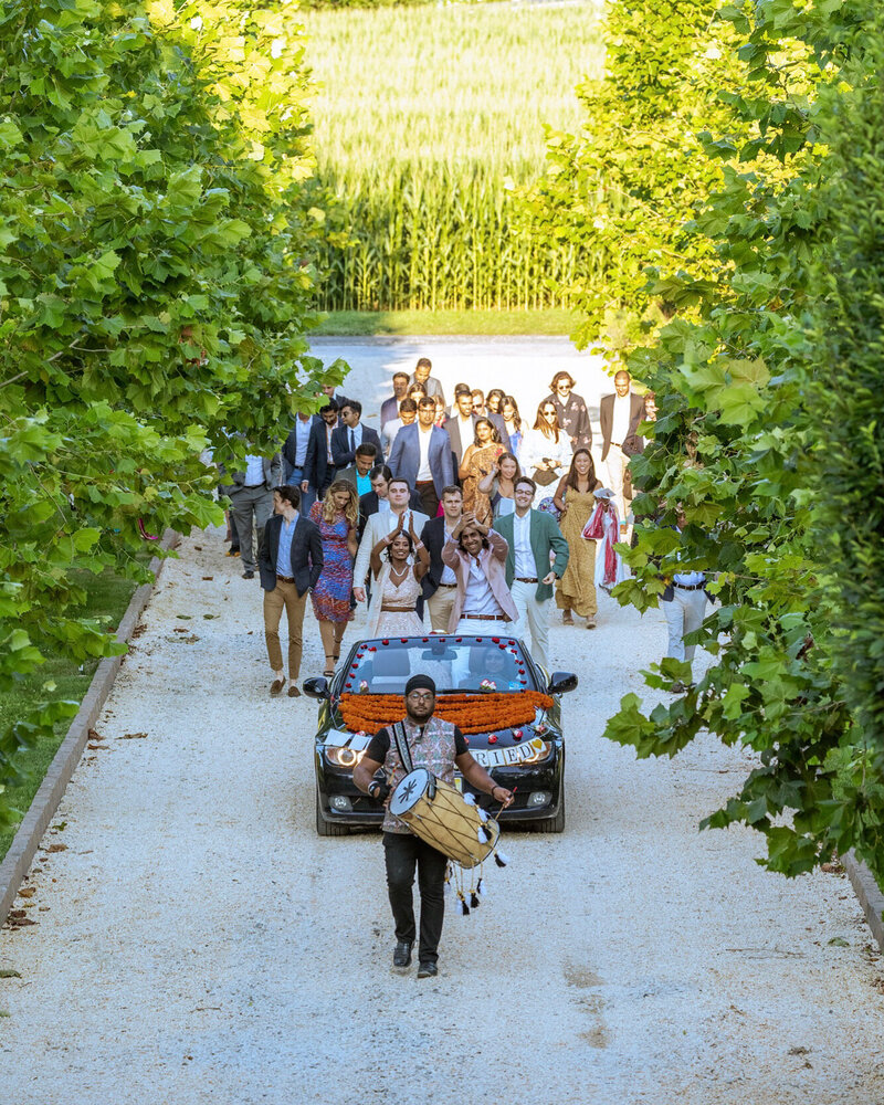 A grand wedding celebration at a private estate in New Jersey, with a lively wedding procession following a decorated car down a scenic gravel path.