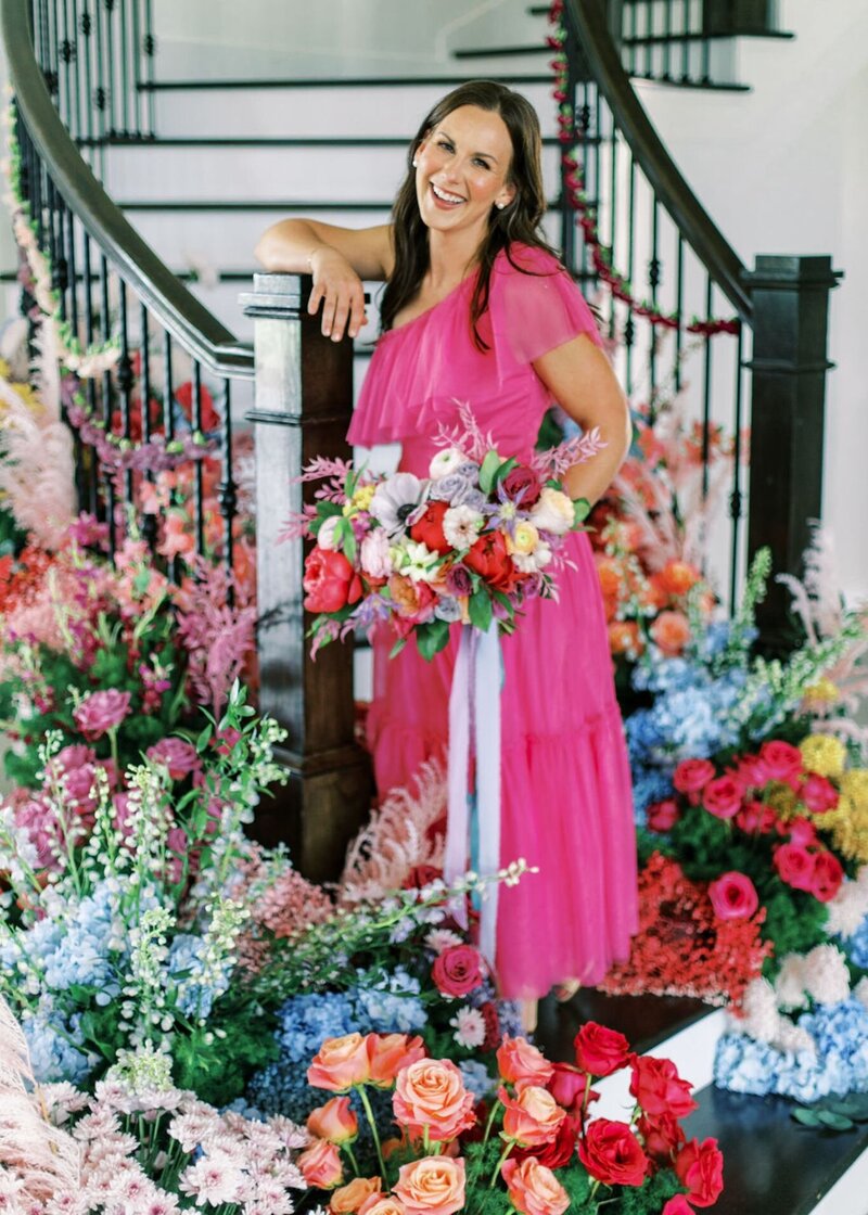 Tamara Menges standing by staircase surrounded by vibrant, colorful floral arrangements.