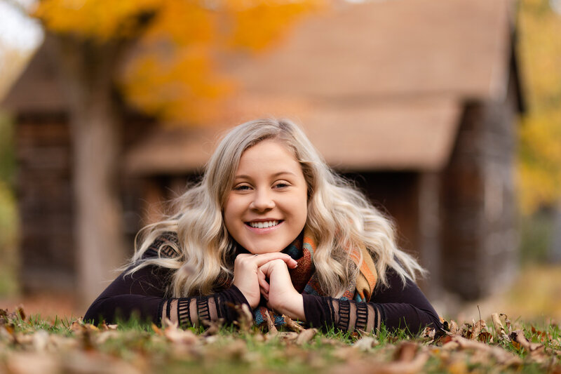 Senior girl laying in a pile of fall leaves smiling at the camera with an old cabin in the background