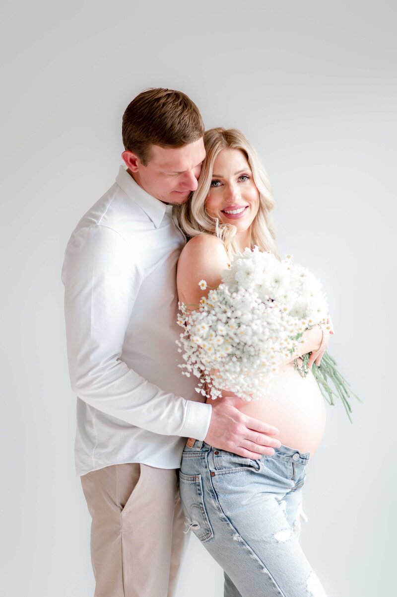 A pregnant woman holds white flowers over her chest and bare belly showing. Her husband stands behind her holding her hip.