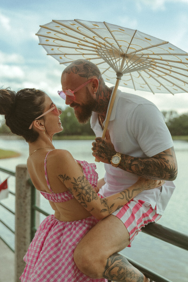 Tattooed couple laughing and holding a parasol in matching pink outfits during a lakeside photoshoot