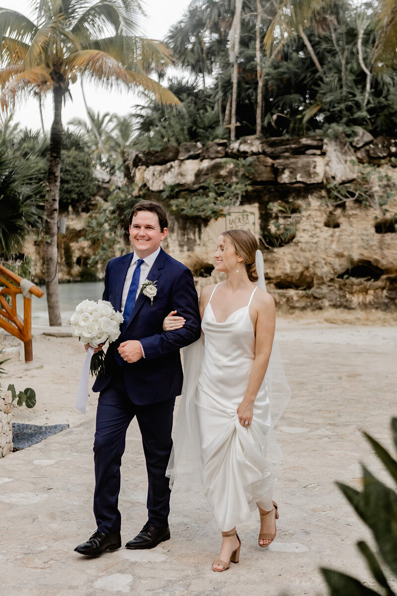 A bride and groom walk arm in arm, smiling after their wedding ceremony. The bride wears a satin gown and holds a bouquet of white roses, while the groom is dressed in a navy blue suit. They are outdoors near a rocky backdrop