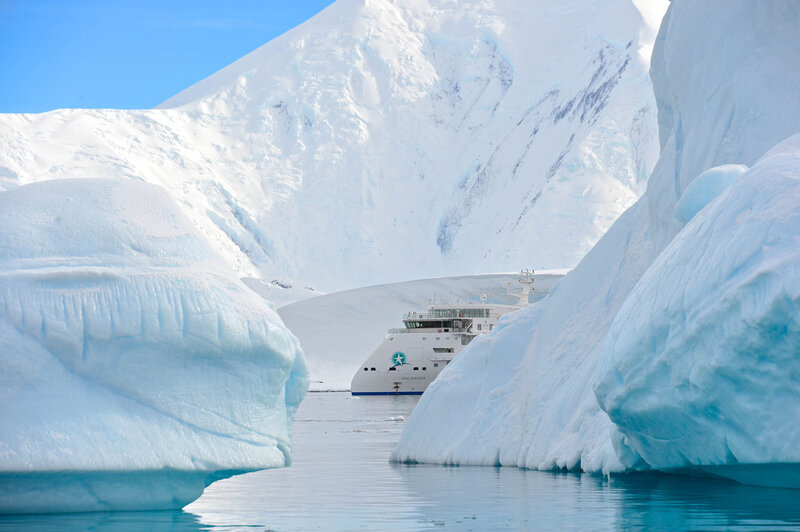 A large ship sailing between massive icebergs in calm, icy waters with snow-covered mountains in the background.
