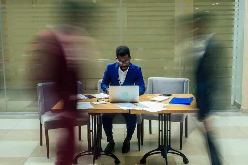 Focused businessman in blue suit working on laptop at a cluttered desk while people walk by in a busy office.