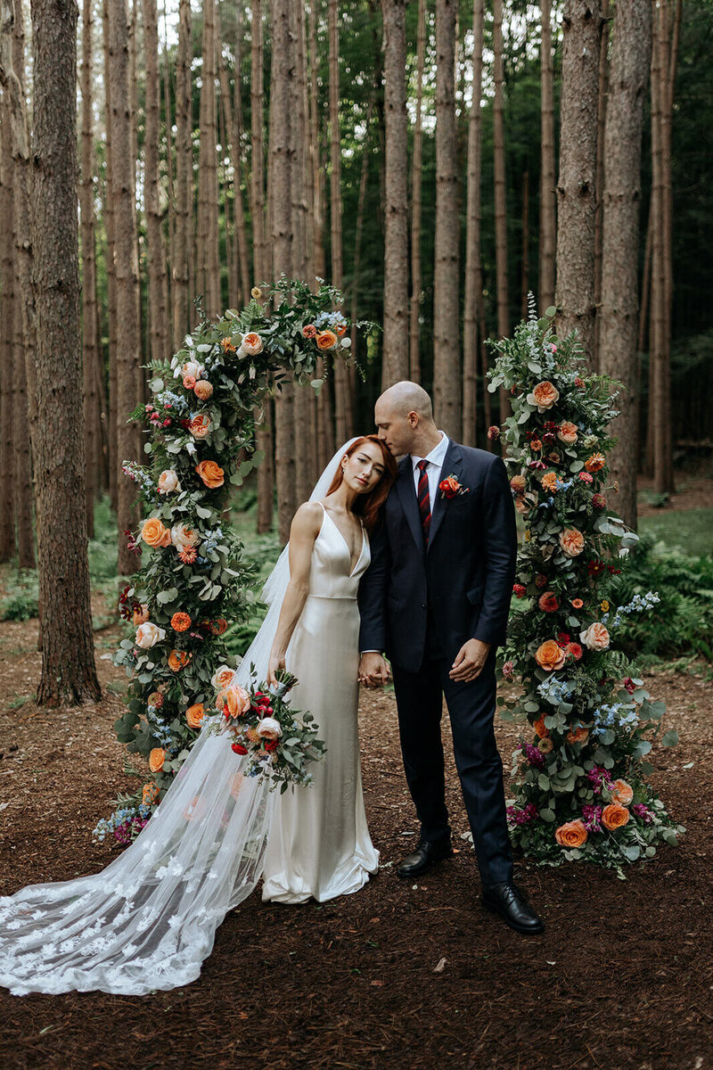A bride and groom stand alone together in th woods before a modern freestanding floral wedding arch.
