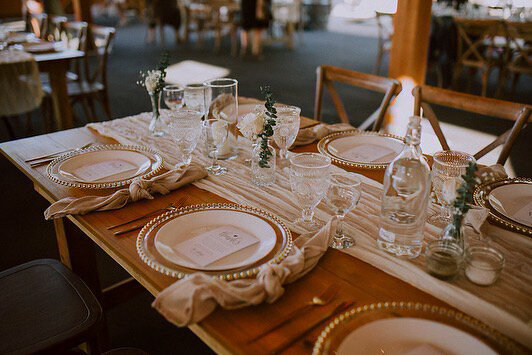 A rustic wedding table adorned with gold-rimmed chargers, crystal glassware, beige napkins, and greenery accents on a wooden table, creating a warm and sophisticated ambiance.