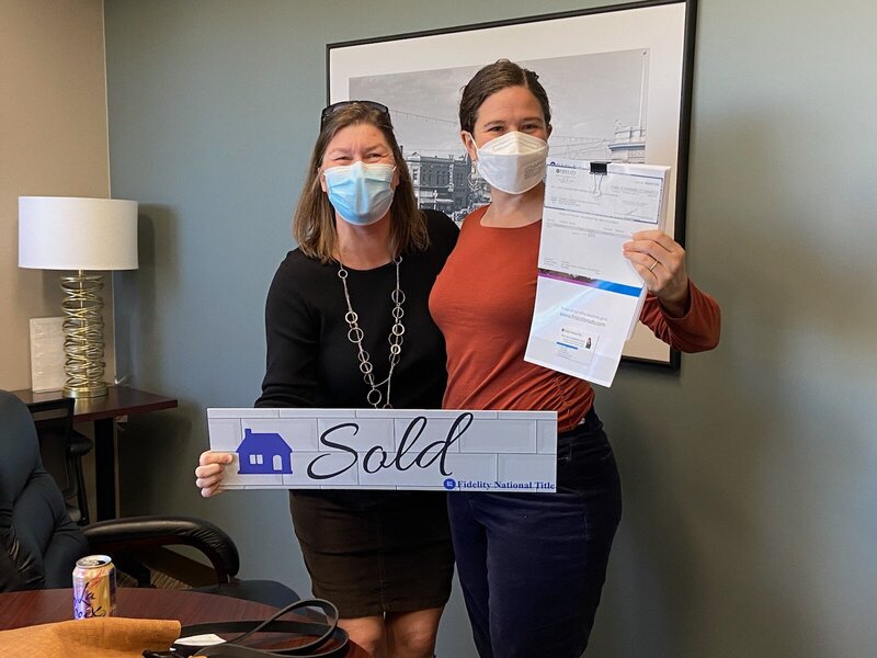 Photo of Mindy, left, and Libby, right at a closing. Mindy is holding a sold sign, little is holding title paperwork