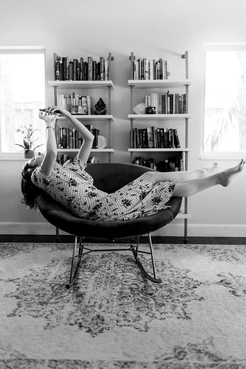 A woman lounging in a chair, holding her phone up while smiling, with bookshelves in the background