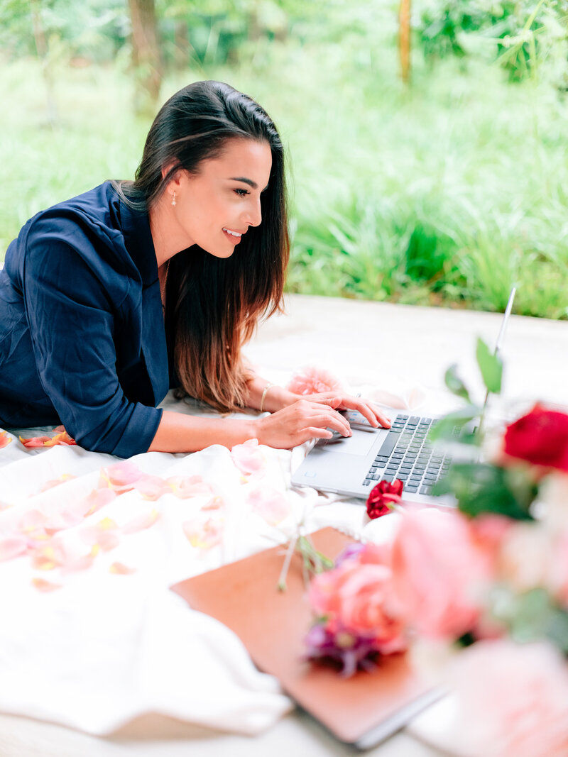 Planner lying on a blanket outdoors, typing on a laptop with scattered rose petals and flowers nearby.